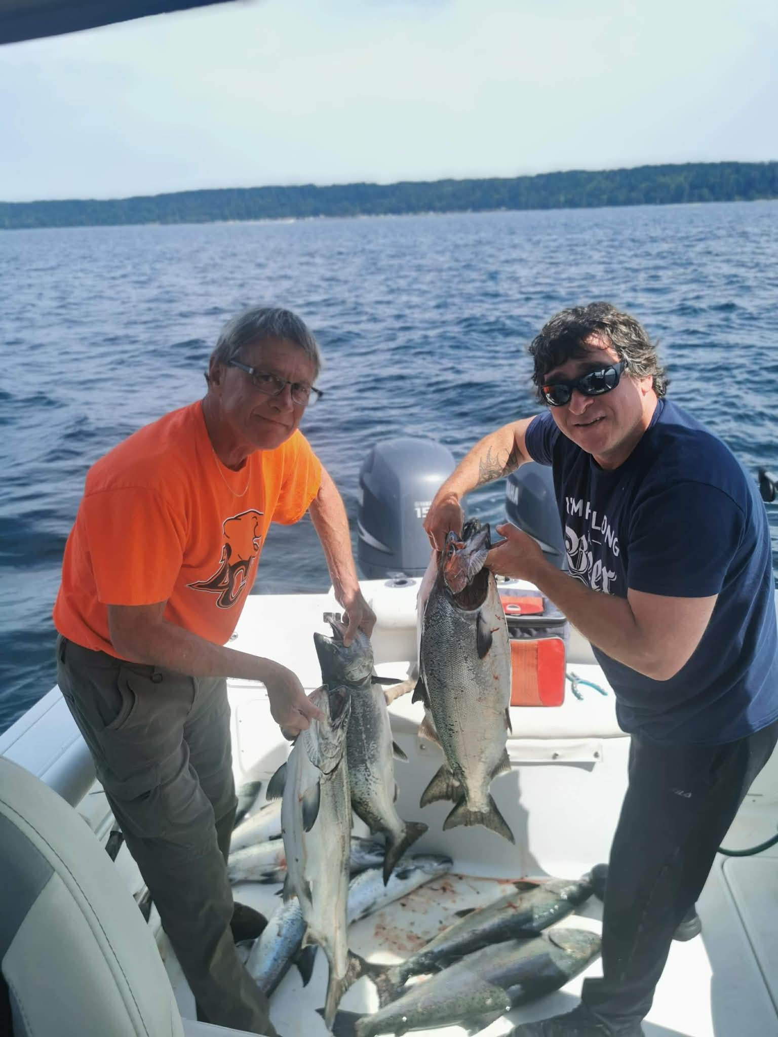 Douglas poses with a man as they hold multiple large fish they caught. They are smiling and standing on the deck of a boat with calm waters and cloudy skies visible in the background.